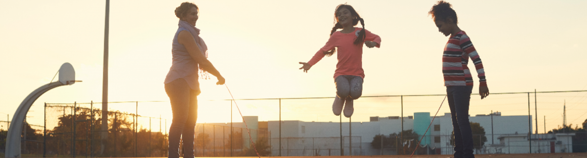 Two girls and their mom play jump rope on an outdoor basketball court as the sun sets behind them.